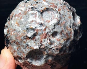 May include: A spherical, hand-held stone sculpture with a rough, textured surface. The stone features a mix of gray, black, and reddish-brown colors, with several small, circular indentations. The overall appearance resembles a miniature planet or celestial body.
