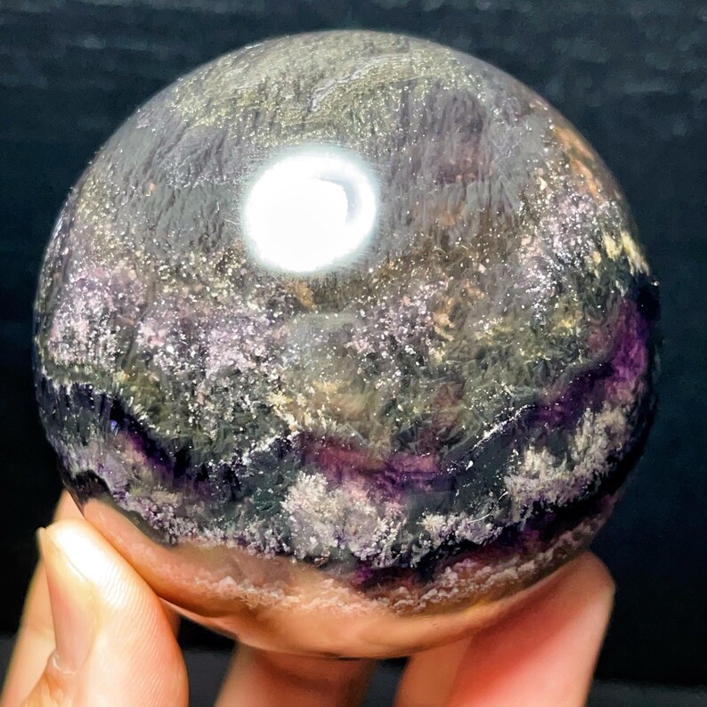 May include: A polished fluorite sphere, displaying a blend of purple, green, and grey hues. The sphere has a smooth, reflective surface with a natural, textured appearance. The crystal ball is held in a hand.