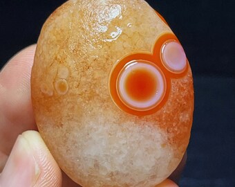 May include: A smooth, light brown agate stone with a unique pattern of concentric circles in shades of orange, white, and pink.