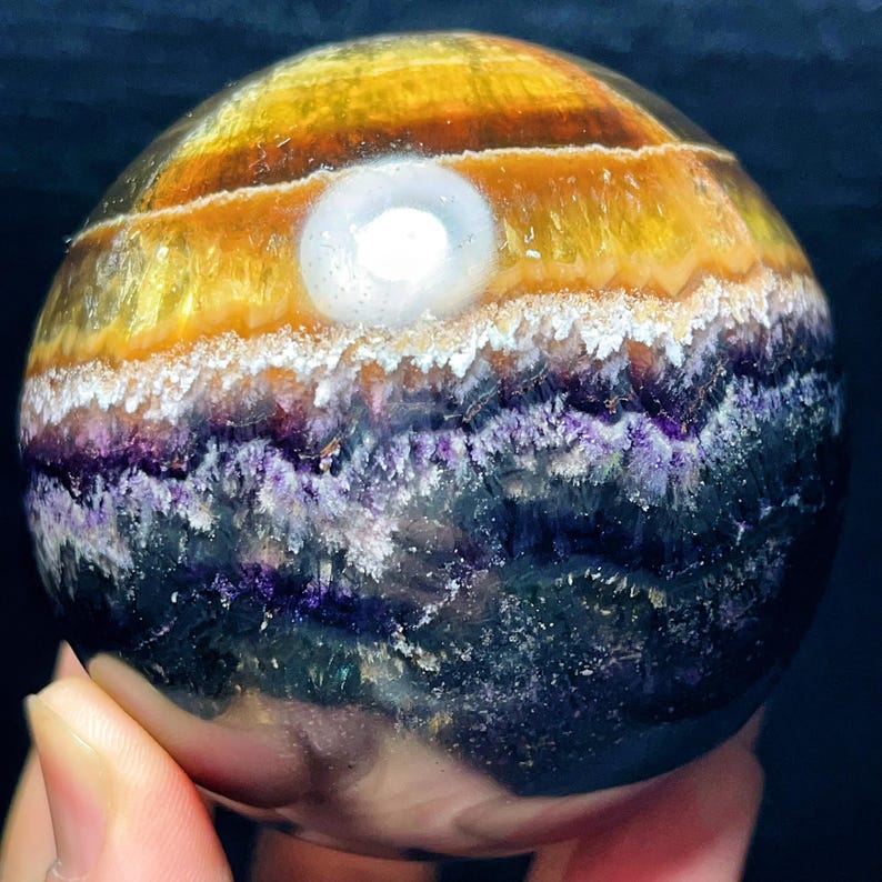 May include: A spherical gemstone with bands of colour. The top section is yellow and orange, transitioning to white, purple, and deep blue at the bottom. A bright, white, circular inclusion is visible near the top.