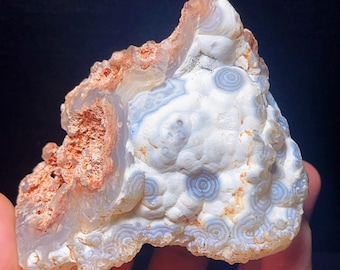 May include: A natural, raw agate specimen with a mix of colors. The stone features a white base with blue, brown, and orange patterns. The agate has a rough, textured surface and a unique, organic shape.