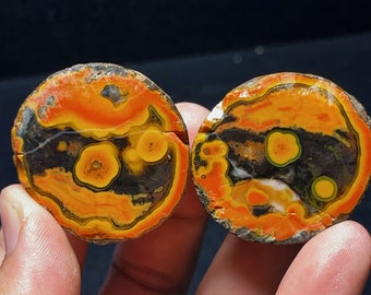 May include: Two round slices of agate with a black, orange, and yellow pattern. The agate has a unique, swirling pattern with a variety of colors.