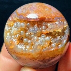 以下が含まれることがあります： 半透明で明るい色のブドウ状の結晶が中央に水平な帯状に広がる、磨かれた球状の石。球体の上部と下部は、オレンジ、茶色、落ち着いたピンクの色合いをした滑らかで不透明な層で構成されています。