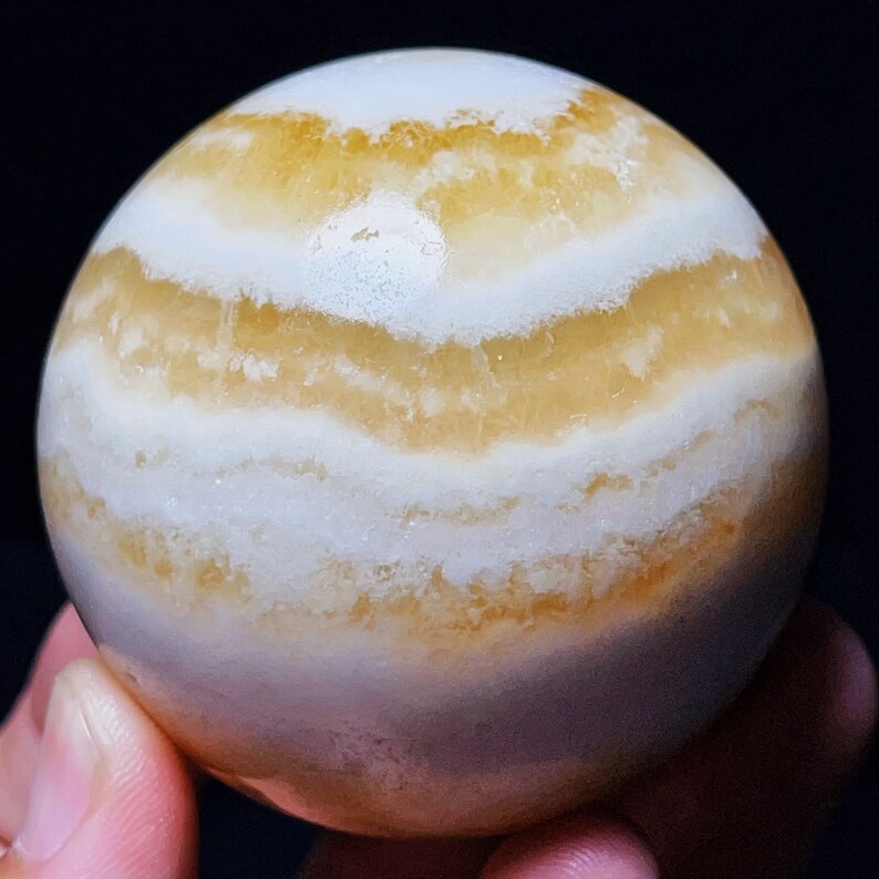 May include: A smooth, white and yellow banded agate sphere. The sphere has a polished finish and is a natural stone.