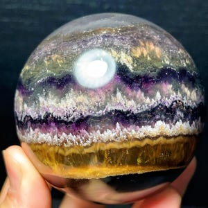 May include: A fluorite sphere, displaying layers of purple, yellow, and white. The gemstone is translucent, with a bright, circular inclusion. The polished ball is held in a hand, highlighting its smooth, spherical shape. The sphere's diameter is approximately 7 cm.