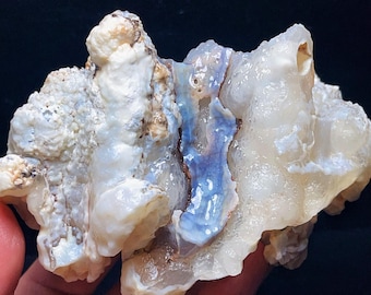 May include: A natural, raw opal specimen with a mix of colors. The opal features a creamy white base with patches of blue and brown. The rough, textured surface of the stone is visible.