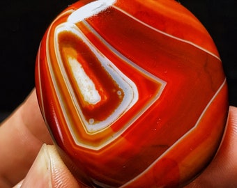 Puede incluir: Una piedra de ágata roja con bandas blancas y marrones. La piedra es lisa y pulida, y tiene una forma natural y orgánica.