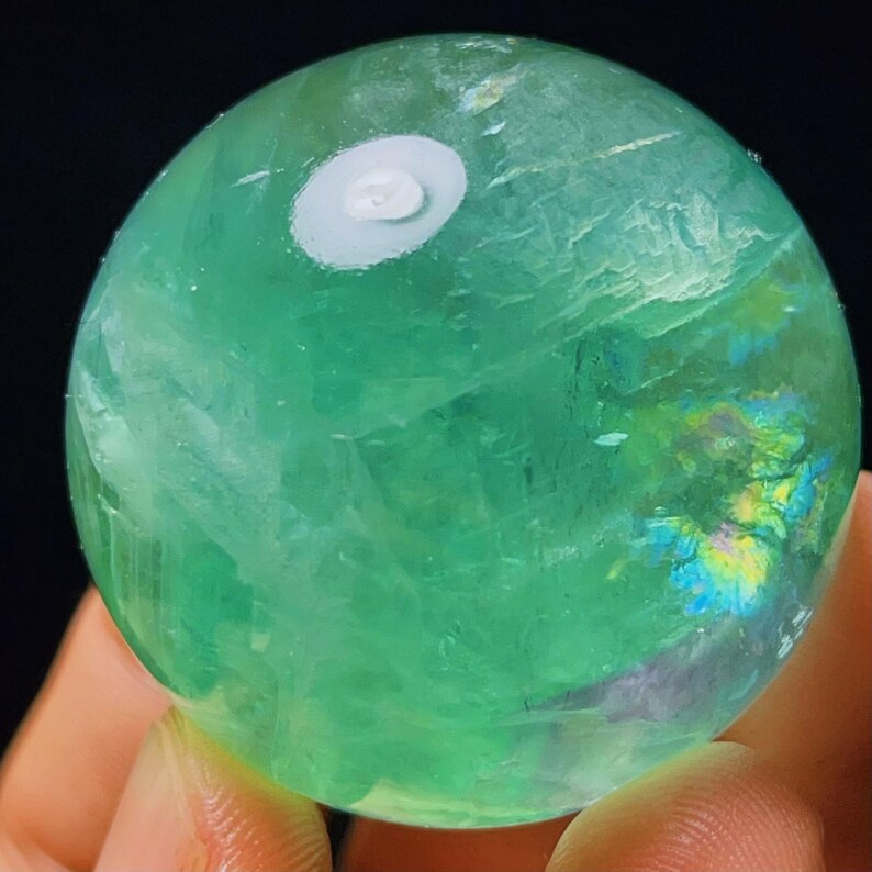 May include: A green fluorite sphere with iridescent accents. The sphere is polished and smooth, with a white base.