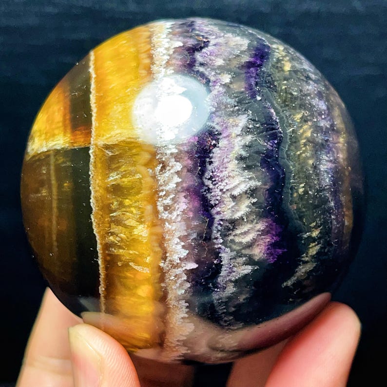 May include: A polished fluorite sphere, displaying bands of yellow, purple, and clear crystal. The gemstone ball is held in a hand, with the colours blending in a natural pattern. The sphere is a decorative item.