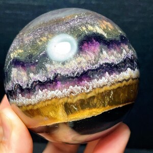 May include: A fluorite sphere, polished to a smooth finish, displaying layers of purple, yellow, and white. The crystal ball has a translucent quality with a bright, white inclusion. The sphere is held in a hand.