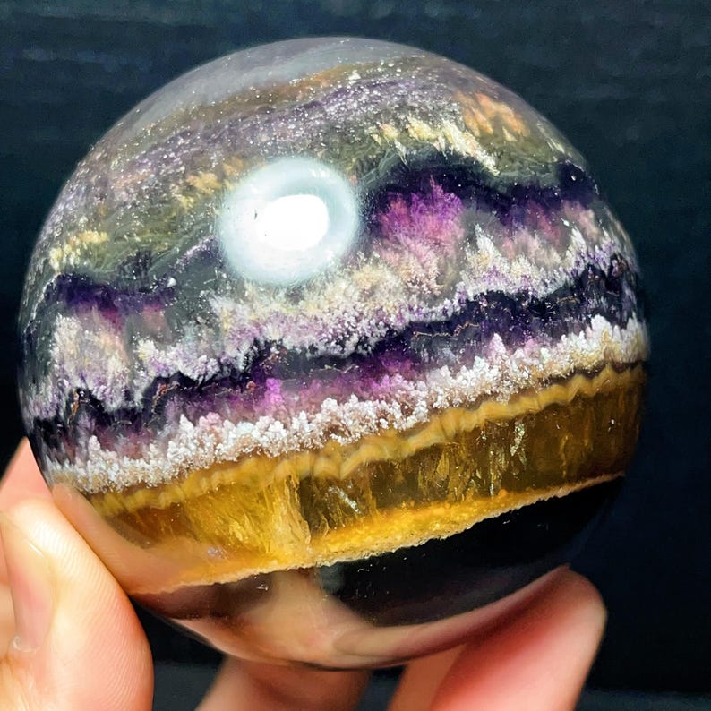 May include: A fluorite sphere, polished to a smooth finish, displaying layers of purple, yellow, and white. The crystal ball has a translucent quality with a bright, white inclusion. The sphere is held in a hand.
