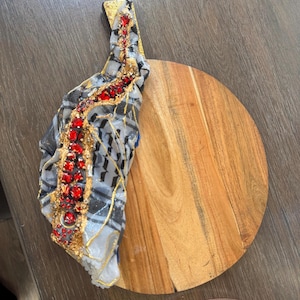 Könnte beinhalten: Ein rundes Holzschneidebrett mit einem dekorativen Griff. Der Griff ist mit goldenen Akzenten und roten Edelsteinen verziert. Ein gemusterter Stoff ist über dem Griff drapiert und verleiht dem Küchenutensil Eleganz.