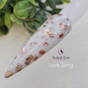 May include: A nail swatch with a white base and embedded gold and copper-colored flakes. The text "Love Song" is displayed below the Soleil Fox logo, indicating a nail dip powder product.