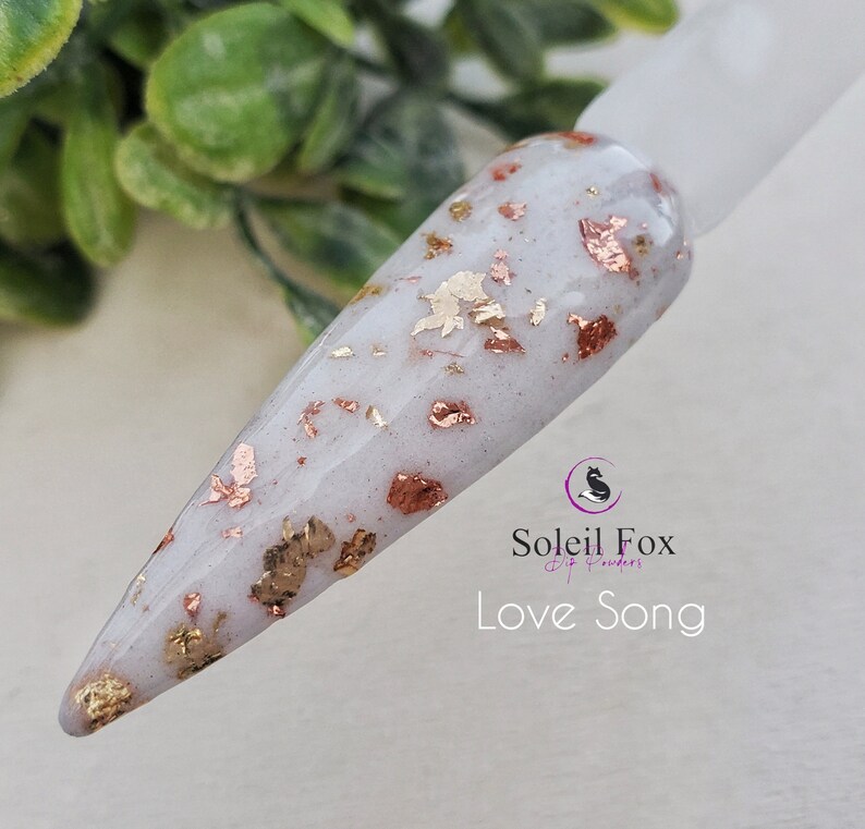 May include: A nail swatch with a white base and embedded gold and copper-colored flakes. The text "Love Song" is displayed below the Soleil Fox logo, indicating a nail dip powder product.
