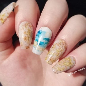 May include: Close-up of a hand with long, coffin-shaped nails. The nails feature a beach-themed design with palm trees and waves, along with gold foil accents. The nails are painted in a neutral beige color.