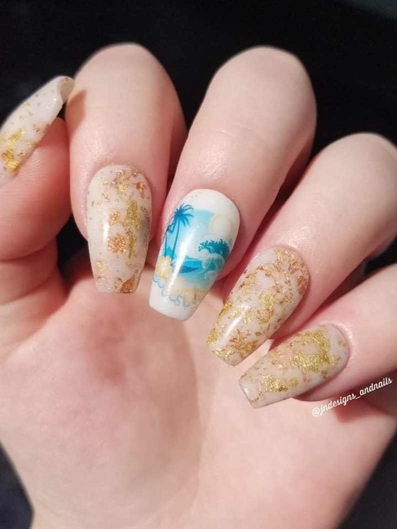 May include: Close-up of a hand with long, coffin-shaped nails. The nails feature a beach-themed design with palm trees and waves, along with gold foil accents. The nails are painted in a neutral beige color.