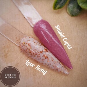 May include: Two nail dip powder samples on clear sticks. One is a shimmery mauve color labeled "Stupid Cupid." The other is a white base with copper flakes, labeled "Love Song." The Soleil Fox Dip Powders logo is in the lower left.