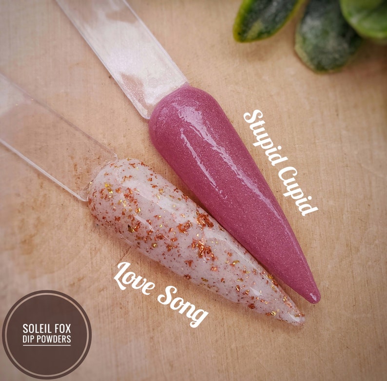 May include: Two nail dip powder samples on clear sticks. One is a shimmery mauve color labeled "Stupid Cupid." The other is a white base with copper flakes, labeled "Love Song." The Soleil Fox Dip Powders logo is in the lower left.
