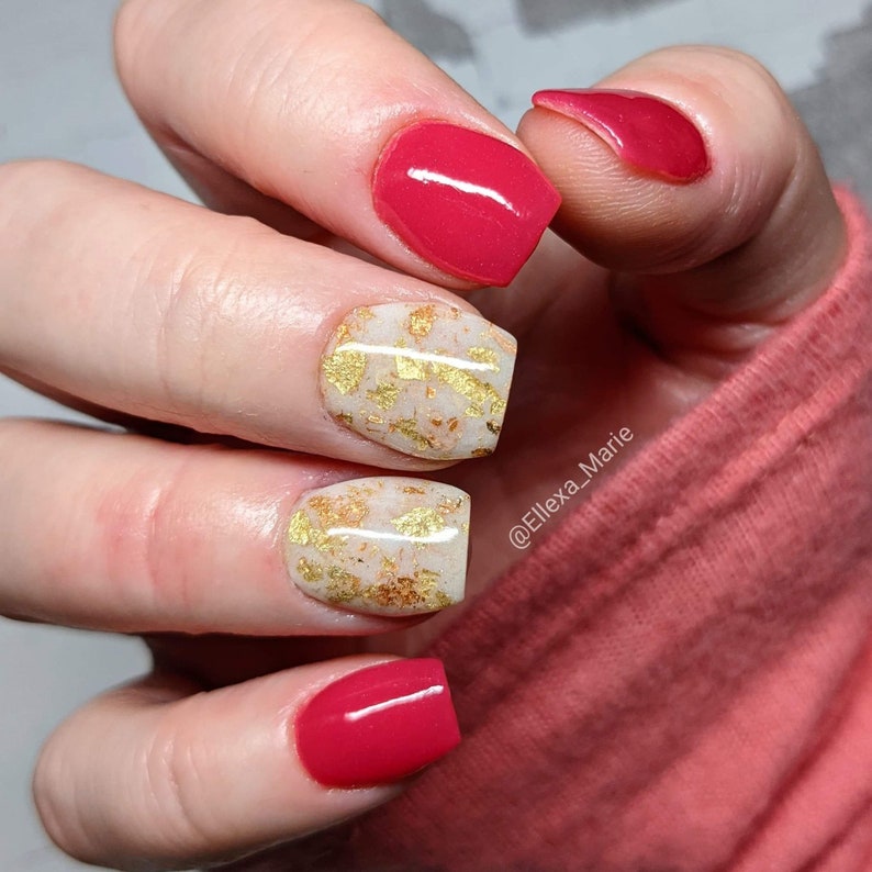 May include: Close-up of a hand with manicured nails. The nails are painted in a vibrant red color and a neutral beige with gold leaf accents. The manicure is a square shape. The image is well-lit and shows the details of the nail art.