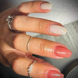 May include: Close-up of a hand with manicured nails. The nails are painted in a light pink and white with gold flecks. The hand is adorned with several silver rings, including a large square-cut diamond ring.