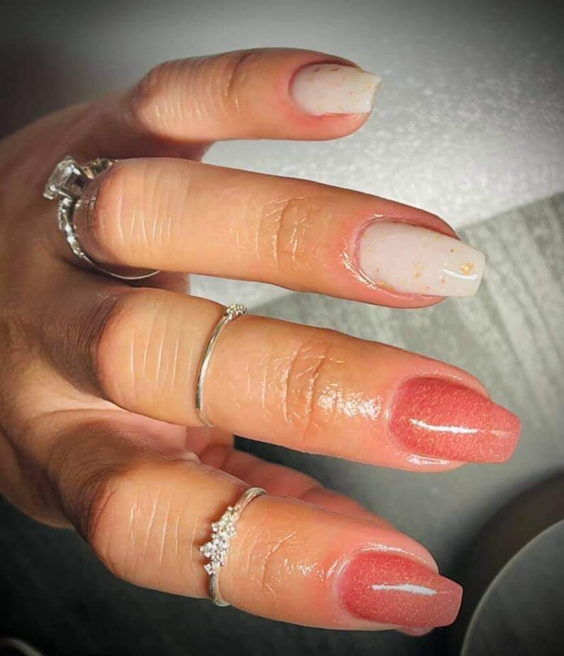 May include: Close-up of a hand with manicured nails. The nails are painted in a light pink and white with gold flecks. The hand is adorned with several silver rings, including a large square-cut diamond ring.