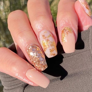 May include: A hand with long, square-shaped nails. The nails are painted in a variety of styles, including a nude base with gold foil accents and a glittery gold design. The text "@Josie.does.nails" is visible on the image.