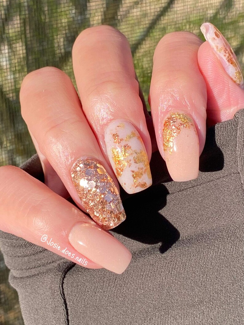 May include: A hand with long, square-shaped nails. The nails are painted in a variety of styles, including a nude base with gold foil accents and a glittery gold design. The text "@Josie.does.nails" is visible on the image.