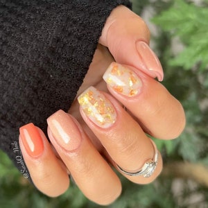 May include: Close-up of a hand with manicured nails. The nails feature a variety of designs, including solid peach, clear with gold flakes, and a vibrant coral color. A silver ring is visible on one finger. The text "@SarahQ_Nails" is on the image.