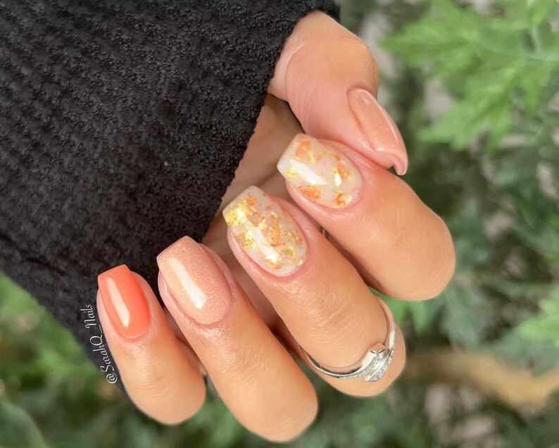 May include: Close-up of a hand with manicured nails. The nails feature a variety of designs, including solid peach, clear with gold flakes, and a vibrant coral color. A silver ring is visible on one finger. The text "@SarahQ_Nails" is on the image.