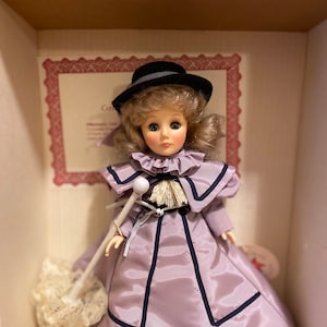 May include: A porcelain doll wearing a lavender dress with black trim, a black hat, and holding a white lace parasol. The doll has blonde hair and blue eyes.