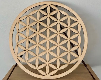 Könnte beinhalten: Ein Wandbehang aus Holz in Form einer Lebensblume. Das komplizierte Design ist aus dem Holz geschnitten und wird mit einer Schnur an einem Regal befestigt.