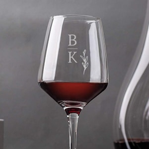 Elegant personalized wine glass with initials 'BK' etched above a simple floral design, filled with red wine, with a decanter and red apples in the background, on a textured grey surface.