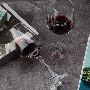 Two custom-engraved wine glasses with names 'Ashley' and 'Sarah' filled with red wine, on a grey marble countertop, with a book and decorative items in the soft-focus background.