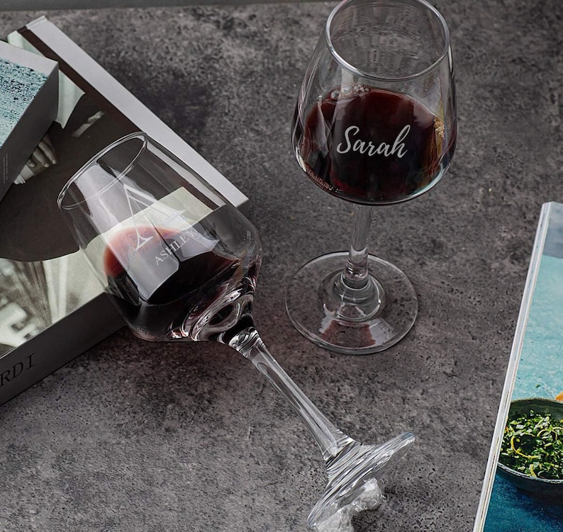 Two custom-engraved wine glasses with names 'Ashley' and 'Sarah' filled with red wine, on a grey marble countertop, with a book and decorative items in the soft-focus background.