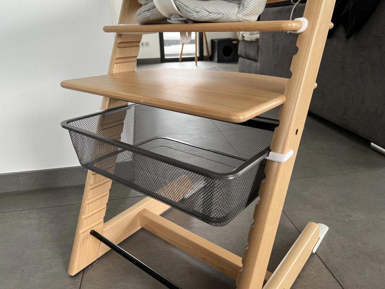 Puede incluir: Una silla alta de madera con una cesta de alambre de metal negro debajo del estante superior. La silla tiene clips de pl&aacute;stico blanco en el lateral.