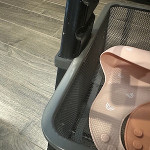 Puede incluir: Una cesta de alambre de metal negro con un babero de silicona rosa con un patr&oacute;n de arco&iacute;ris blanco en su interior. El babero tiene una secci&oacute;n de silicona marr&oacute;n con un patr&oacute;n de plumas.