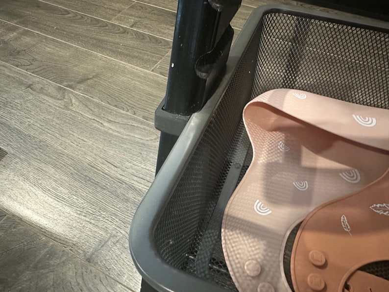 Puede incluir: Una cesta de alambre de metal negro con un babero de silicona rosa con un patr&oacute;n de arco&iacute;ris blanco en su interior. El babero tiene una secci&oacute;n de silicona marr&oacute;n con un patr&oacute;n de plumas.