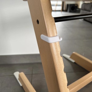 Puede incluir: Primer plano de una silla alta de madera con una correa de seguridad de pl&aacute;stico blanco. La silla tiene un acabado de madera clara y una serie de ranuras a lo largo del lado.