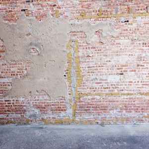 May include: A weathered brick wall with patches of peeling plaster and a concrete floor in front of it.