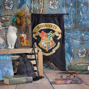 Pode incluir: Uma caixa de madeira rústica com uma pilha de livros de Harry Potter, um chapéu de bruxa preto, uma varinha de madeira e duas corujas brancas. Uma faixa com o brasão da casa de Hogwarts está pendurada no fundo. A cena é ambientada contra uma parede texturizada azul e marrom.