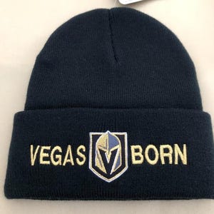 May include: A navy blue knit beanie with the words "VEGAS BORN" embroidered in gold. A gold, navy, and white team logo is centered on the front. A tag with a black cat logo is attached.