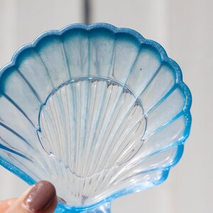 Puede incluir: Un plato de vidrio azul y blanco en forma de concha con una superficie texturizada. El plato tiene forma de concha y tiene un borde azul.