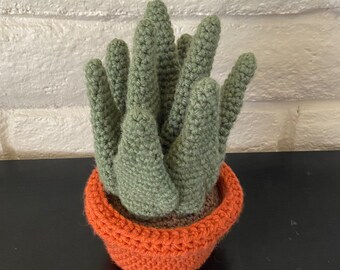 May include: A handmade crochet cactus in a bright orange pot. The cactus is a soft green color and has multiple upright stems. The pot is a vibrant orange, and the base of the cactus is a brown color.