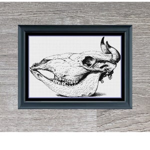 May include: Black and white cross-stitch pattern of a bull skull, framed in black, on a grey wood background. The title "Bull Skull" and dimensions "274 x 182" are printed below the frame.