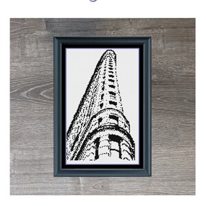 May include: A black and white cross-stitch pattern of the Flatiron Building in New York City. The pattern is framed and displayed against a wood background. The text "Mini Flatiron Building" is below the image. The dimensions are 95 x 155 stitches and it uses 1 DMC colour.