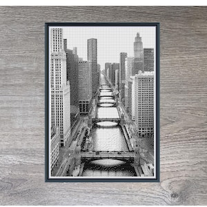 Peut inclure: Une œuvre d'art en point de croix encadrée en noir et blanc représentant une scène de ville avec de grands immeubles bordant une rivière. L'œuvre est présentée sur une surface en bois. Le texte "Stitching Jules Design" est visible en haut.