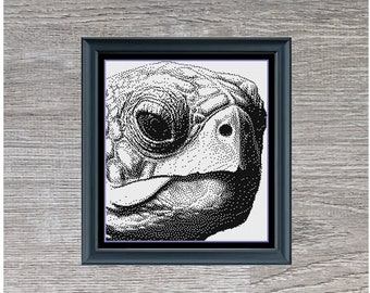 May include: Black and white cross-stitch pattern of a tortoise's head. The pattern is framed in a black frame and set against a woodgrain background. The text "Tortoise Monochrome" and "188 x 210" is below the image.