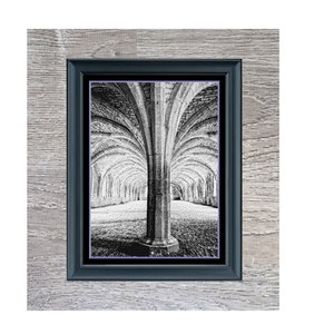 Op de afbeelding: Zwart-wit print van een gewelfde stenen gang, getiteld "The Cellarium", in een donkerblauwe lijst. De afbeelding staat op een lichtgrijze houten achtergrond. De print toont een lange, gewelfde gang.