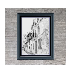 May include: Black and white sketch of a narrow street in Greece with buildings on either side. The image is framed in a black frame and set against a woodgrain background. The text "Greek Side Street" is below the image.