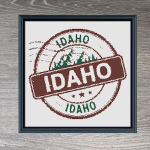 Può includere: Un disegno a punto croce incorniciato con un timbro circolare con la parola "IDAHO" in un banner rettangolare. Il timbro include un grafico di montagna e alberi, con il testo "IDAHO" ripetuto. Il design è nei toni del marrone, verde e bianco.